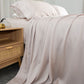 Dusty pink bed sheets draped, to one side, showcasing the sheet texture as well as pilows popped up on the bed next to a bedside table with ornaments.