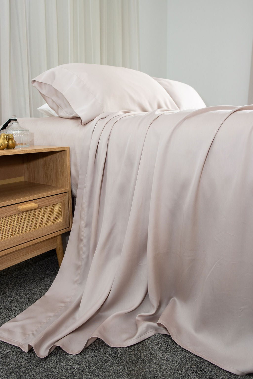 Dusty pink bed sheets draped, to one side, showcasing the sheet texture as well as pilows popped up on the bed next to a bedside table with ornaments.